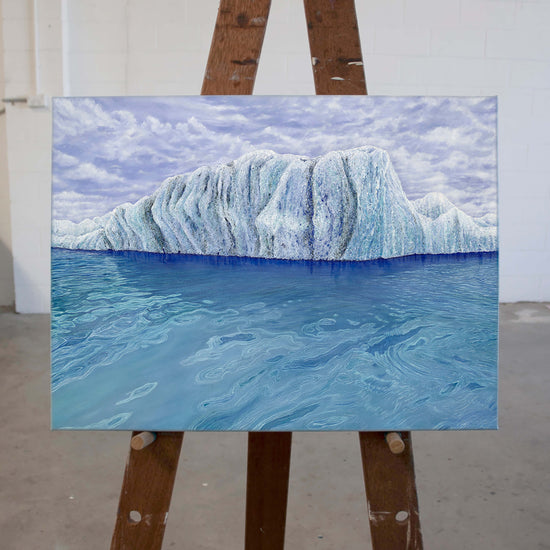 Original art, titled Ammonite, painted in oil on canvas, by Australian artist, Hayley Byron. The painting is a depiction of an iceberg, with clouds in the background, and water in the foreground. This image features the artwork on an easel.