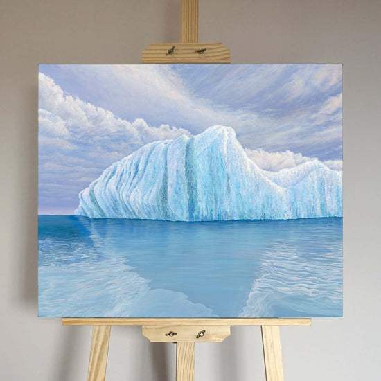 Original art, titled Ametrine, painted in oil on linen, by Australian artist, Hayley Byron. The painting is a depiction of an iceberg, with clouds in the background, and water in the foreground. This image features the artwork on an easel in an interior room.
