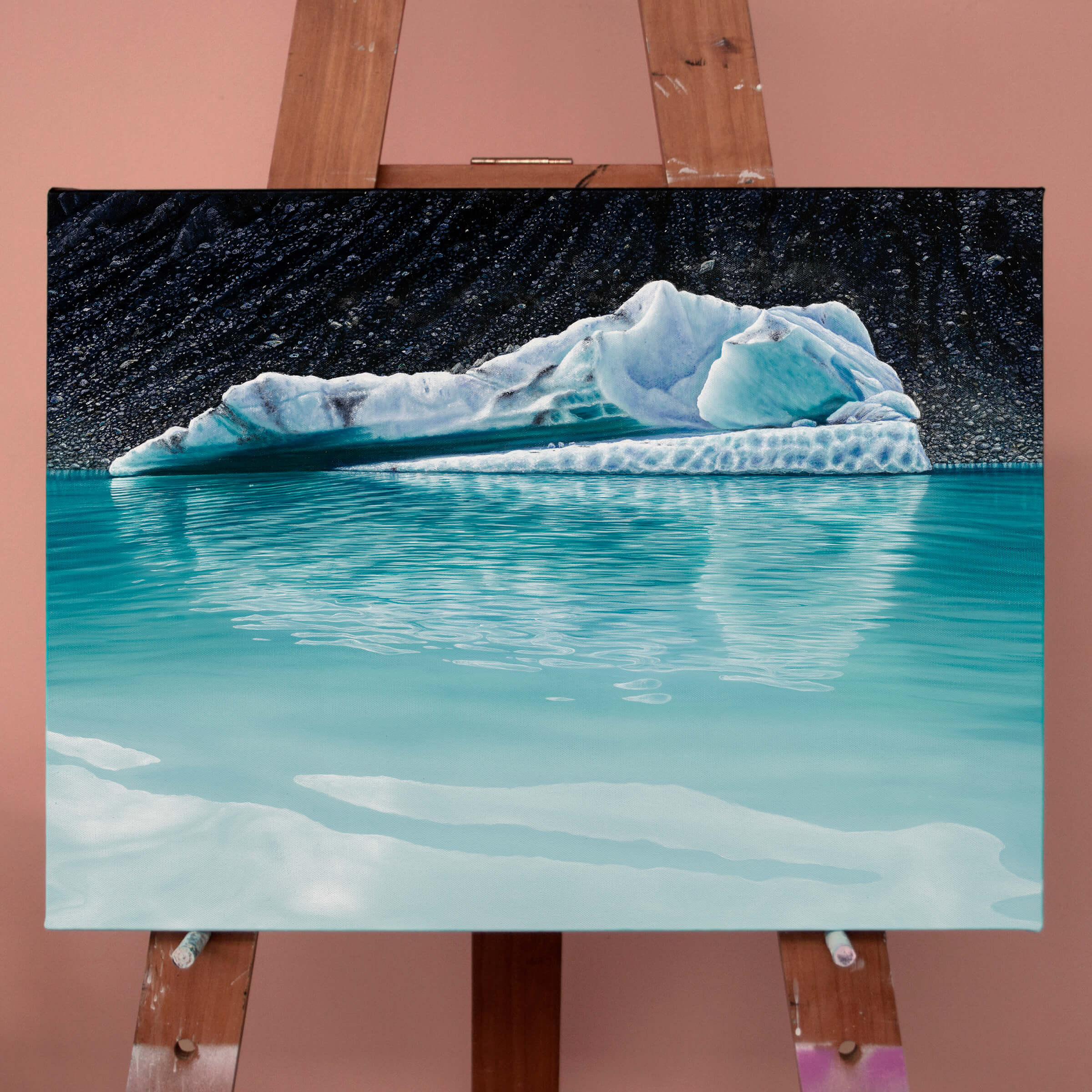 Original art, titled Chrysocolla, painted in oil on canvas, by Australian artist, Hayley Byron. The painting is a depiction of an iceberg, with glacial till in the background, and water in the foreground. This image features the artwork on an easel.