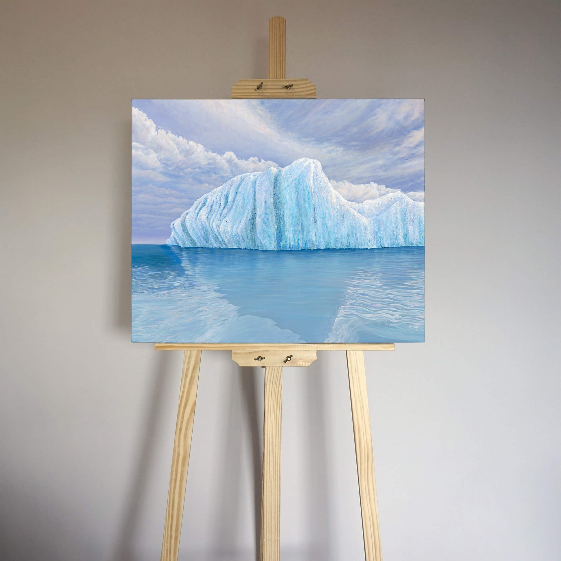 Original art, titled Ametrine, painted in oil on linen, by Australian artist, Hayley Byron. The painting is a depiction of an iceberg, with clouds in the background, and water in the foreground. This image features the artwork on an easel in an interior room.