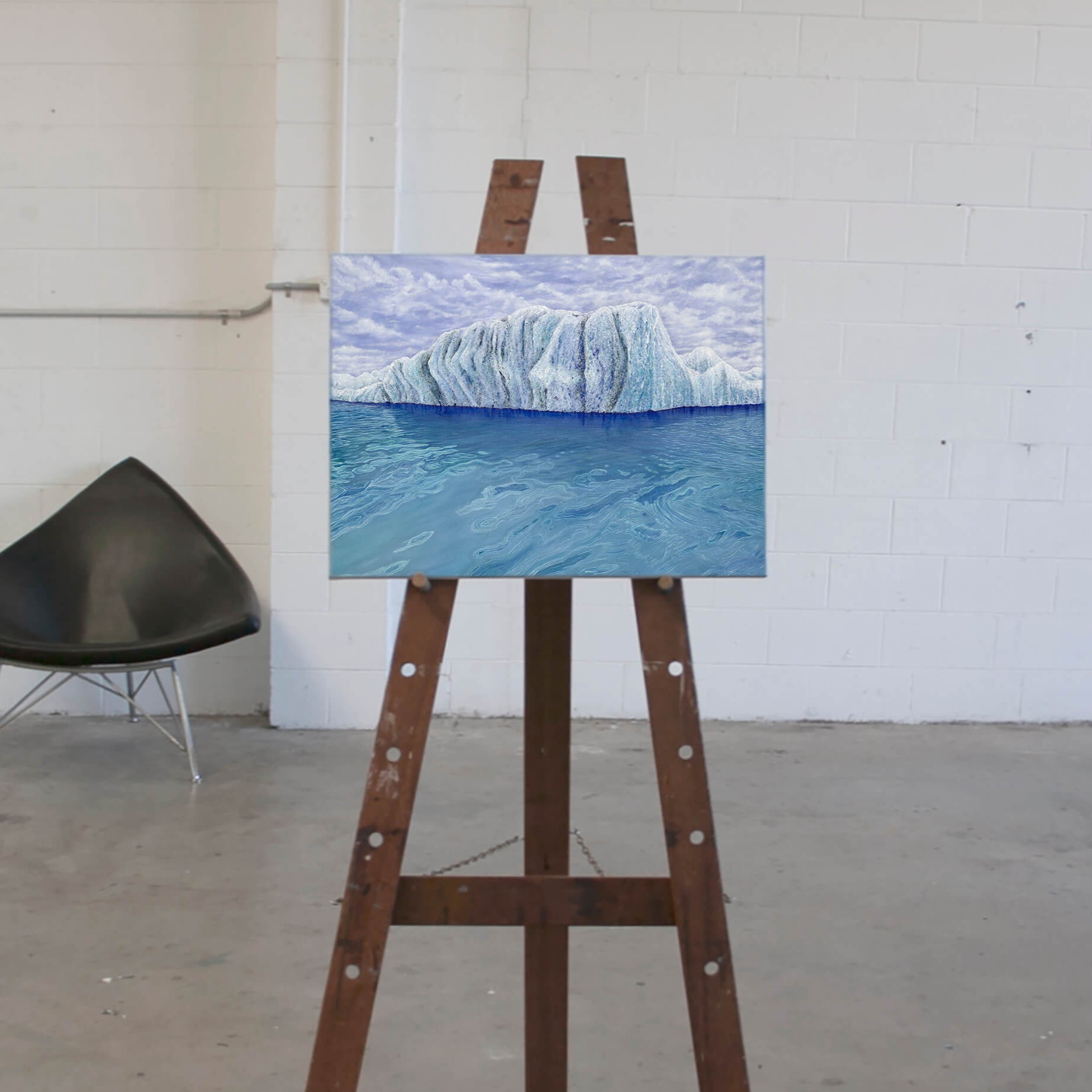 Original art, titled Ammonite, painted in oil on canvas, by Australian artist, Hayley Byron. The painting is a depiction of an iceberg, with clouds in the background, and water in the foreground. This image features the artwork on an easel in an art studio.