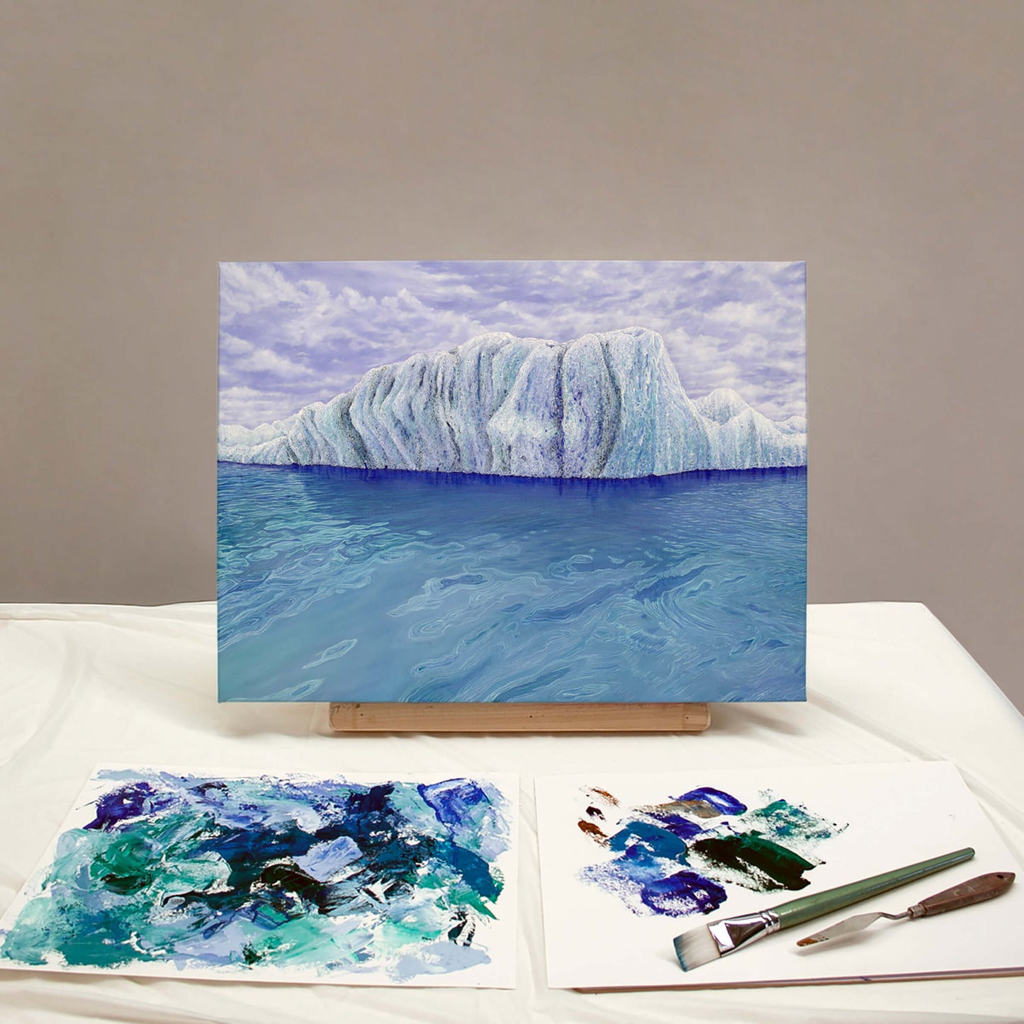 Original art, titled Ammonite, painted in oil on canvas, by Australian artist, Hayley Byron. The painting is a depiction of an iceberg, with clouds in the background, and water in the foreground. This image features the artwork on an easel in an art studio, next to a paint palette and paintbrushes.
