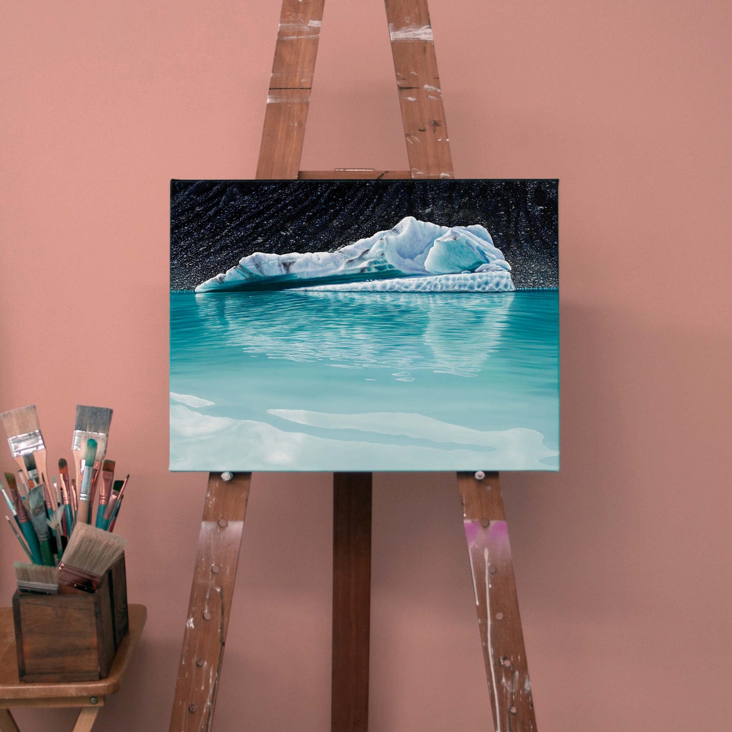 Original art, titled Chrysocolla, painted in oil on canvas, by Australian artist, Hayley Byron. The painting is a depiction of an iceberg, with glacial till in the background, and water in the foreground. This image features the artwork on an easel, next to paintbrushes, in an art studio.