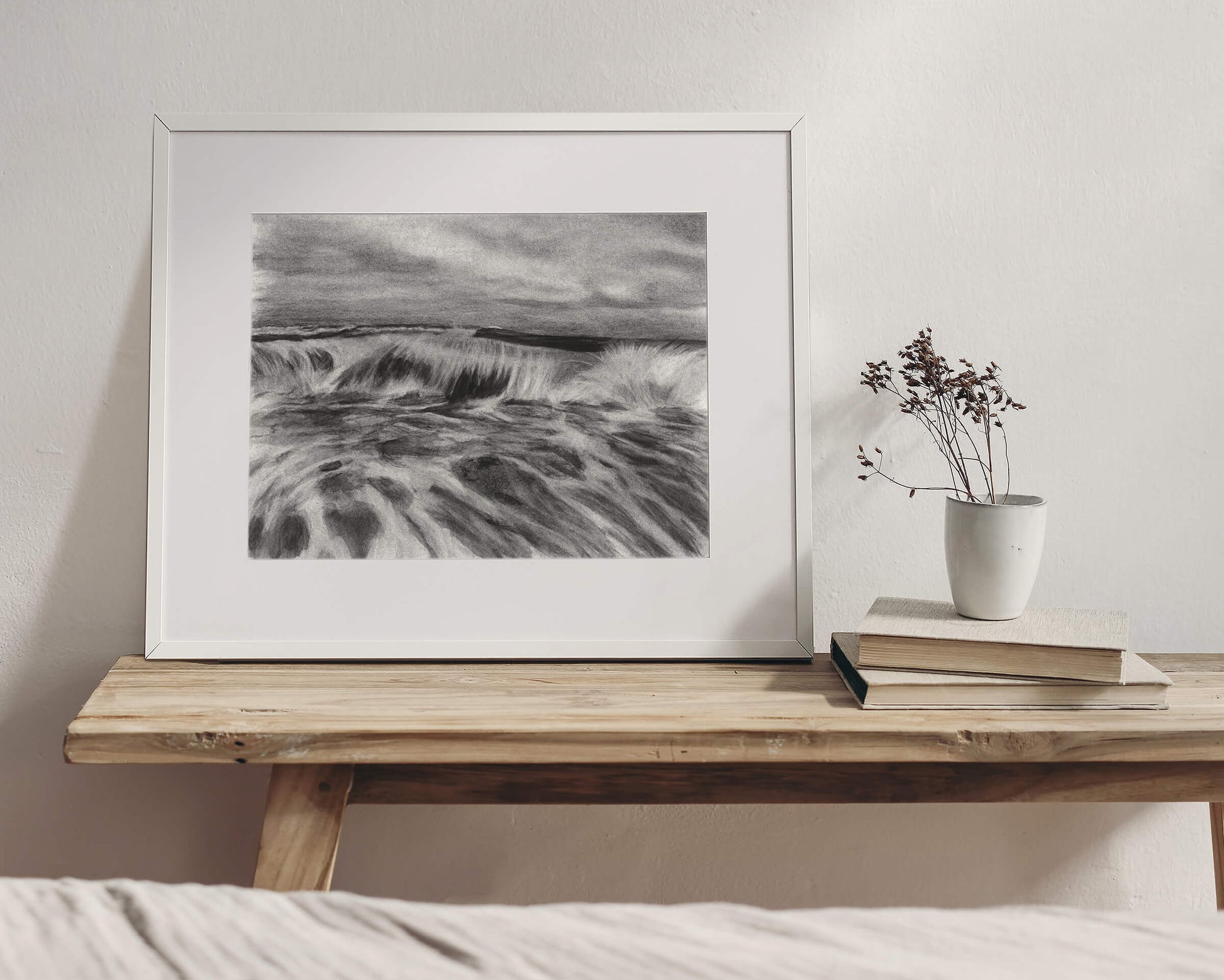 Original art, titled La Mar, hand-drawn with graphite and willow charcoal on fine art paper, by Australian artist, Hayley Byron. The drawing is a depiction of a wave in the ocean. This image features the artwork in-situ, positioned on a table, next to books and a ceramic vase, in an interior bedroom.